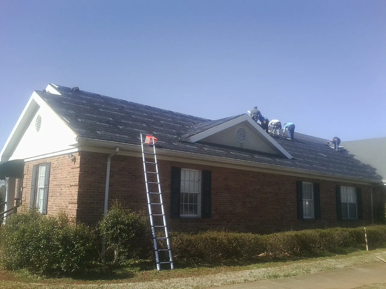 Crew tearing shingles off a brick ranch roof with a ladder set — Emergency Roof Repair work by our crew in Rogers